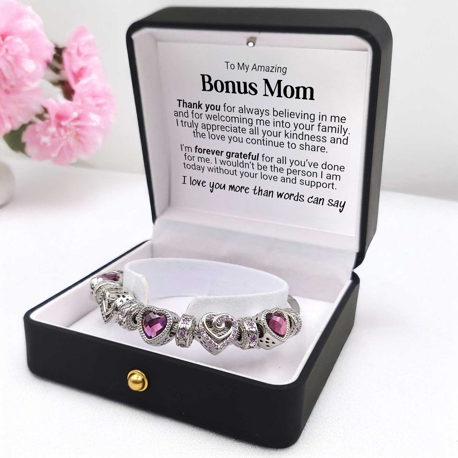Bracelet with heart-shaped charms in a gift box with a message for 'Bonus Mom' on a white background with pink flowers.