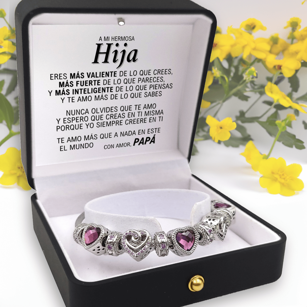 Bracelet with purple heart charms in a black box with a message for daughter in Spanish, surrounded by yellow flowers.