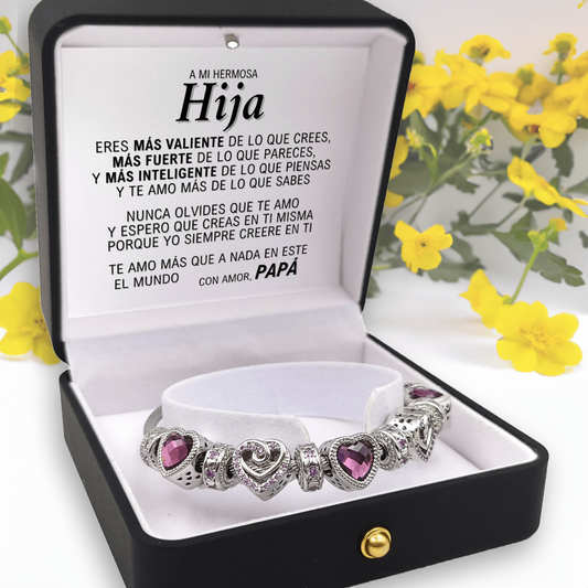 Bracelet with purple heart charms in a black box with a message for daughter in Spanish, surrounded by yellow flowers.