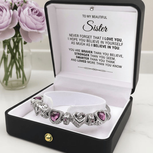 Silver bracelet with purple heart stones in a black box with a message for a sister, next to a vase of purple flowers.