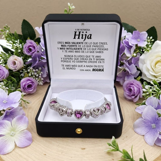 Purple bracelet with heart charms in a black box with Spanish text, surrounded by flowers.